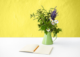 inspiration concept - notebook or sketchbook with pencil and bunch of flowers in green rustic jug on white table over yellow background