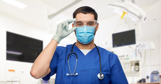 medicine, healthcare and surgery concept - doctor or male nurse in blue uniform, face medical mask for protection from virus disease, goggles and gloves over operating room at hospital background