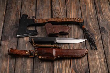 On a wooden background lies an ax in a leather case and a survival knife with a hand sheath, soft...