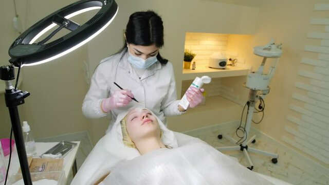 Experienced cosmetician is applying face mask on female face. Girl is lying and relaxing. Her eyes are closed with pleasure