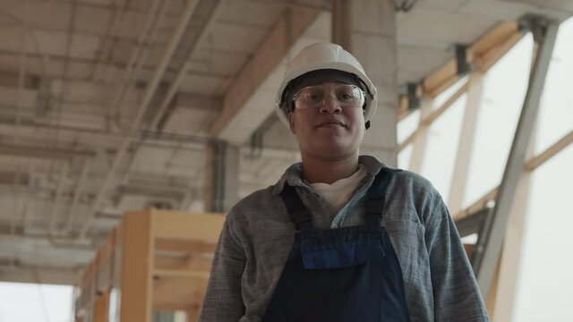 Low angle arc medium point of view of Mixed-Race female builder wearing protective goggles and helmet, standing in house under construction, looking on camera