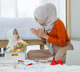 A cute and beautiful Asian Muslim mother playing with her baby daughter during arrange for kid clothes and work on laptop notebook computer.