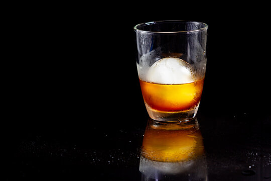 Ice Ball In Glass Of Whisky On A Reflective Black Table.