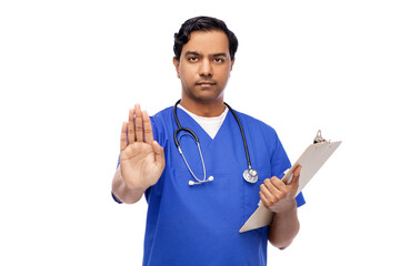 healthcare, profession and medicine concept - indian male doctor in blue uniform with stethoscope and clipboard showing stop gesture over white background