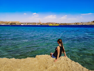Young woman sits on a cliff above the turquoise-blue-azure water of the Red Sea in Sharm El Sheikh (Egypt). Adult girl tourist on a stone coast against the background of an exotic tropical seascape