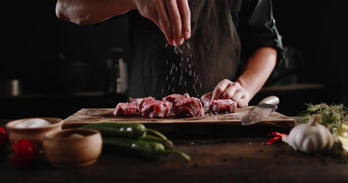 Chef cooks beef steak, sprinkling with salt on a background of professional kitchen, slow motion