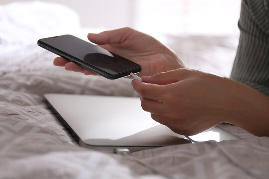 Woman connecting charger cable to smartphone near laptop on bed, closeup