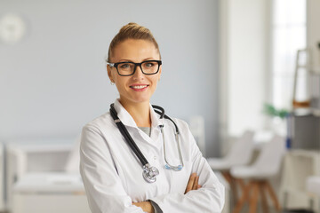 Portrait of friendly doctor or nurse at work. Professional medical staff headshot. Happy young woman in eyeglasses and white lab coat with stethoscope standing arms crossed and smiling at camera