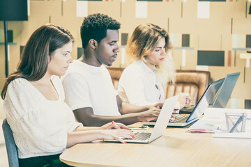 Creative professionals sitting together at table with blueprints and working on project. Row of colleagues typing on laptops. Side view. Coworking space concept