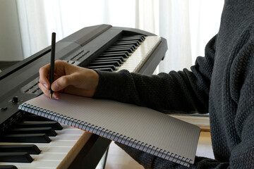 Close up shot of a young music student composing a song in his notebook in front of the piano, in the comfort of his home. Copy space