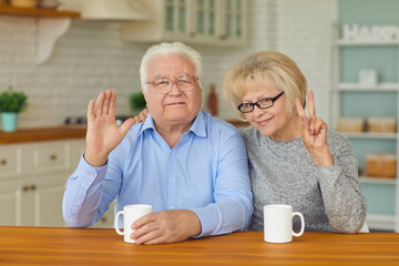Happy elderly couple grandparents sitting together in kitchen with hot drinks, looking at camera, waving and showing peace sign with fingers. Elderly people happy lifestyle, family happiness concept
