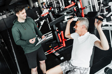 Two men in the gym, one of them a physiotherapist holding a training plan in his hands, the other...