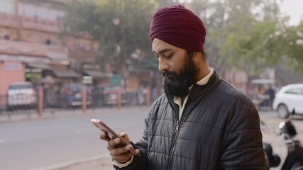 shot of a young Indian office worker man wearing turban on head with long beard using a mobile phone standing by the corner of a street during morning hours