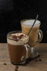 Two glass cups with a cappuccino and an iced chocolate on a wooden table with a black textured background