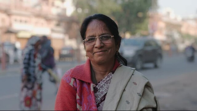 close view shot of a middle aged indian asian common woman wearing specs and traditional saree with smile on face standing at the side of a urban local street and looking at camera 