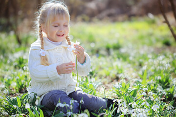 A girl examines a snowdrop flower. A girl in a warm white sweater walks through the spring forest.