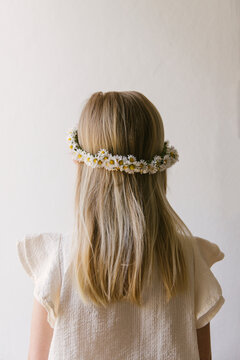 Blonde haired girl wearing a daisy crown seen in front of a empty neutral white background wearing a white summer blouse with her back turned to the camera.