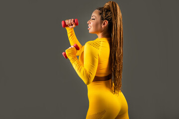 beautiful adult woman in yellow tracksuit on gray background with dumbbells 
