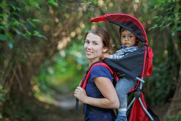 Mother with toddler child in backpack carrier is walking in forest. Tourist is carrying a baby on...