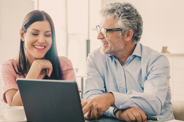 Joyful male and female colleagues of different ages meeting over cup of coffee at co-working, sitting at open laptop, discussing project and laughing. Medium shot. Consulting or cooperation concept