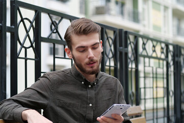 Handsome businessman sits on bench and holds in hand white smartphone