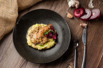 Baked meat and mashed potatoes served with mushrooms, green sauce and lettuce in black bowl. Close-up on a wooden background with a sacking, sliced onion, garlic and silver cutlery by side. Top view.