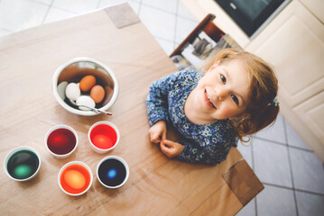 Excited little toddler girl coloring eggs for Easter. Cute happy child looking surprised at colorful colored eggs, celebrating holiday with family. From above