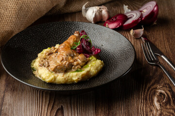 Baked meat and mashed potatoes served with mushrooms, green sauce and lettuce in black bowl. Close-up on a wooden background with a sacking, sliced onion, cloves of garlic and silver cutlery by side.