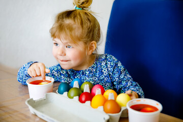 Excited little toddler girl coloring eggs for Easter. Child looking surprised at colored egg hoarding and celebrating catholic and christian holiday with family. Cute kid helping to color, indoors.