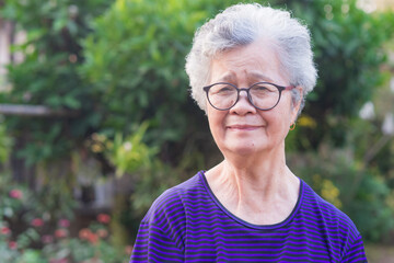 Portrait of a happy senior woman wearing glasses with short gray hair smiling and looking at the camera while standing in a garden. Space for text. Concept of aged people and healthcare