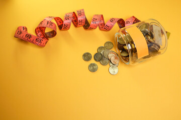 View from above of a group of coins flowing out from the glass jar and measuring tape on the yellow background 