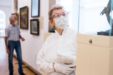 Fototapeta premium elderly European woman in mask protecting against covid examines paintings on display in hall of art museum