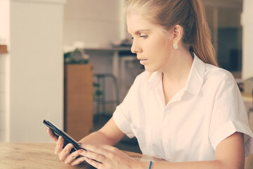 Pensive beautiful blonde woman wearing white shirt, using tablet while sitting at table in co-working space. Medium shot. Digital communication concept