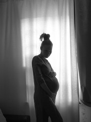 The silhouette of a pregnant girl standing by the window. Black and white photo.