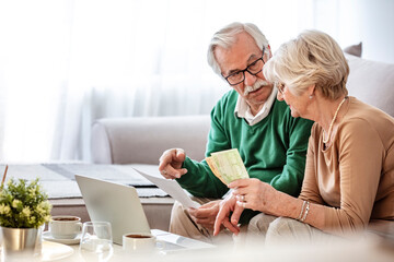 Senior couple sitting at a table at home and going through their household finances using a laptop. Senior couple going through bills while using laptop at home. - obrazy, fototapety, plakaty