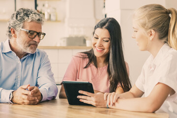 Female agent or manager meeting with couple of young and mature customers, presenting content on tablet. Man and woman looking at screen, smiling, laughing. Consulting or technology concept