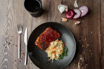 Baked meat served with red sauce, sauerkraut and dill in black bowl. Close-up on a wooden background with a black glass, onion, garlic, mix of pepper seeds and cutlery by side. Top view, copy space.