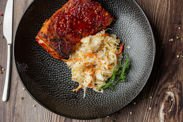 Baked meat served with red sauce, sauerkraut and dill in a black bowl. Top view macro close-up on a wooden background with a mix of pepper seeds and silver cutlery by side.
