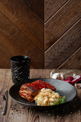 Baked meat served with red sauce, sauerkraut and dill in black bowl. Close-up on wooden background with black glass, sliced onion, cloves of garlic, mix of pepper seeds and a fork by side. Copy space.