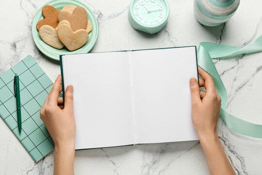 Female hands with blank open book, clock and cookies on light background