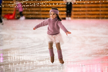 Little girl figure skater in pink sweater is skating on winter evening on an outdoor ice rink lit by garlands © Maria Moroz