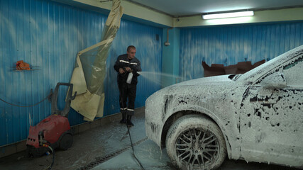 A Caucasian man works in a car wash service. A man in a worker's uniform sprays a car with detergent