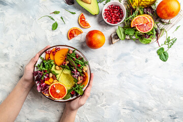 Girls' hands holding vegan, detox Buddha bowl with avocado, persimmon, blood orange, nuts, spinach, arugula and pomegranate on a light background, top view