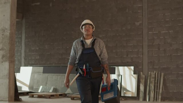 Medium Long Of Mixed-Race Woman-construction Worker Wearing Helmet And Goggles, Carrying Toolbox, Walking Confidently Towards Camera In Building In Progress