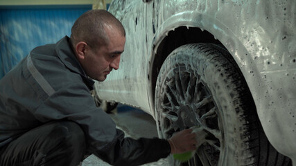 Fototapeta na wymiar A Caucasian man works in a car wash service. A man in a worker's uniform washes the wheel of a car thoroughly