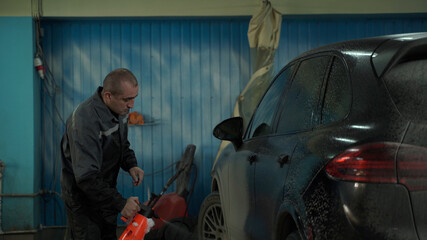 A Caucasian man works in a car wash service. A man in a worker's uniform washes the car thoroughly by spraying it with a water pistol.