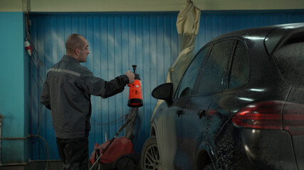 A Caucasian man works in a car wash service. A man in a worker's uniform washes the car thoroughly by spraying it with a water pistol.
