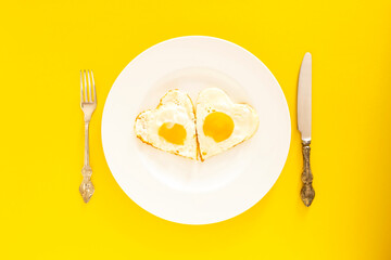 Heart shaped fried eggs in a white plate, silver cutlery on the yellow background. Top view. Copy space.