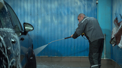 A Caucasian man works in a car wash service. A man in a worker's uniform washes the car thoroughly by spraying it with a water pistol.