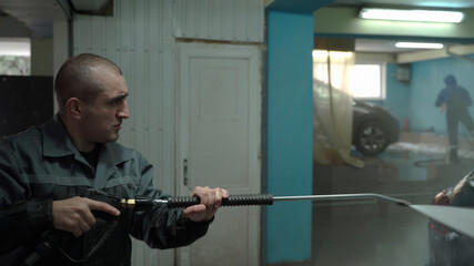 A Caucasian man works in a car wash service. A man in a worker's uniform holds a water gun and washes the car with a jet of water